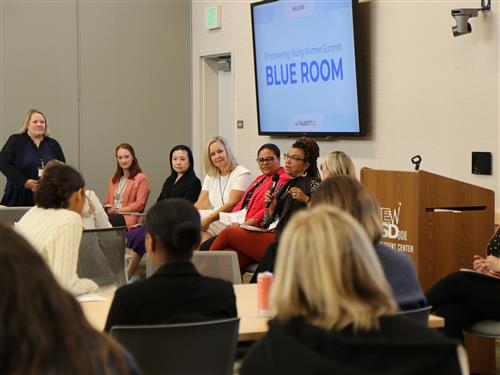 A woman speaking into a microphone on a panel with other women to a group of high school students. 